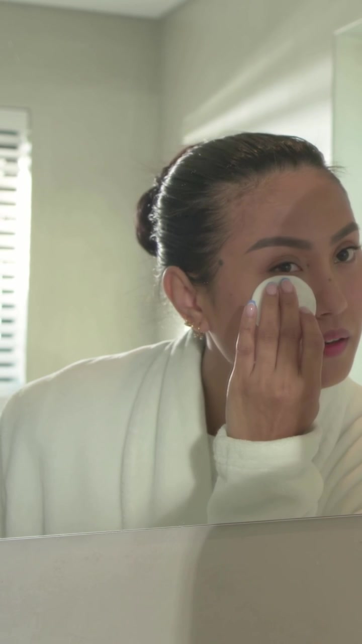 Vertical video: Glancing Asian woman in robe reaching for cotton pad and cleansing face at sink