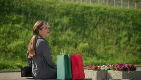 woman approaches bench carrying colorful shopping bags, sits down, and relaxes her left hand on the bench, wind flutters her hair as she adjusts herself, with greenery and flowers in the background