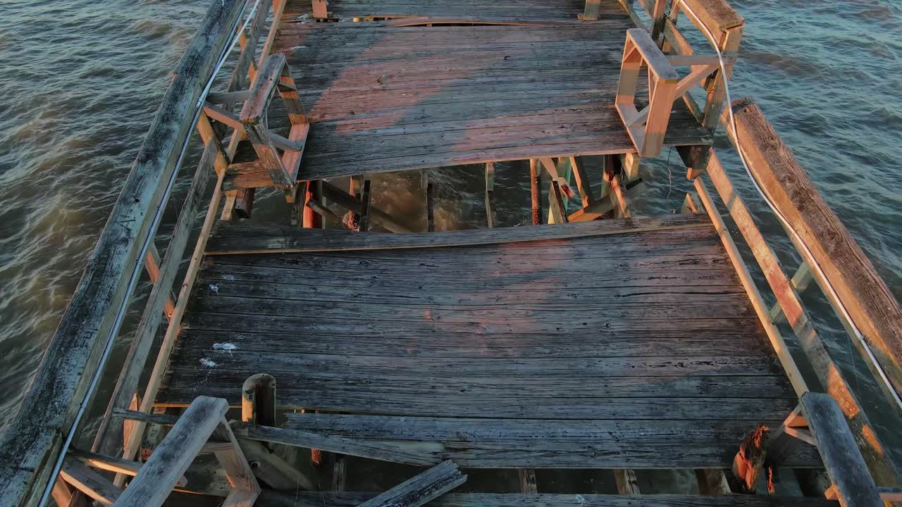 Aerial tilt up shot over broken wooden jetty pier during sunset and sandy beach in background