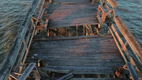 toma aérea inclinada hacia arriba sobre un embarcadero de madera roto durante la puesta de sol y la playa de arena en el fondo