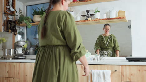 cook in green gown with braided hair walks into kitchen as her front reflects in wall mirror, surrounded by wooden cabinets, potted plants, utensils, mixer, and countertop kitchen tools