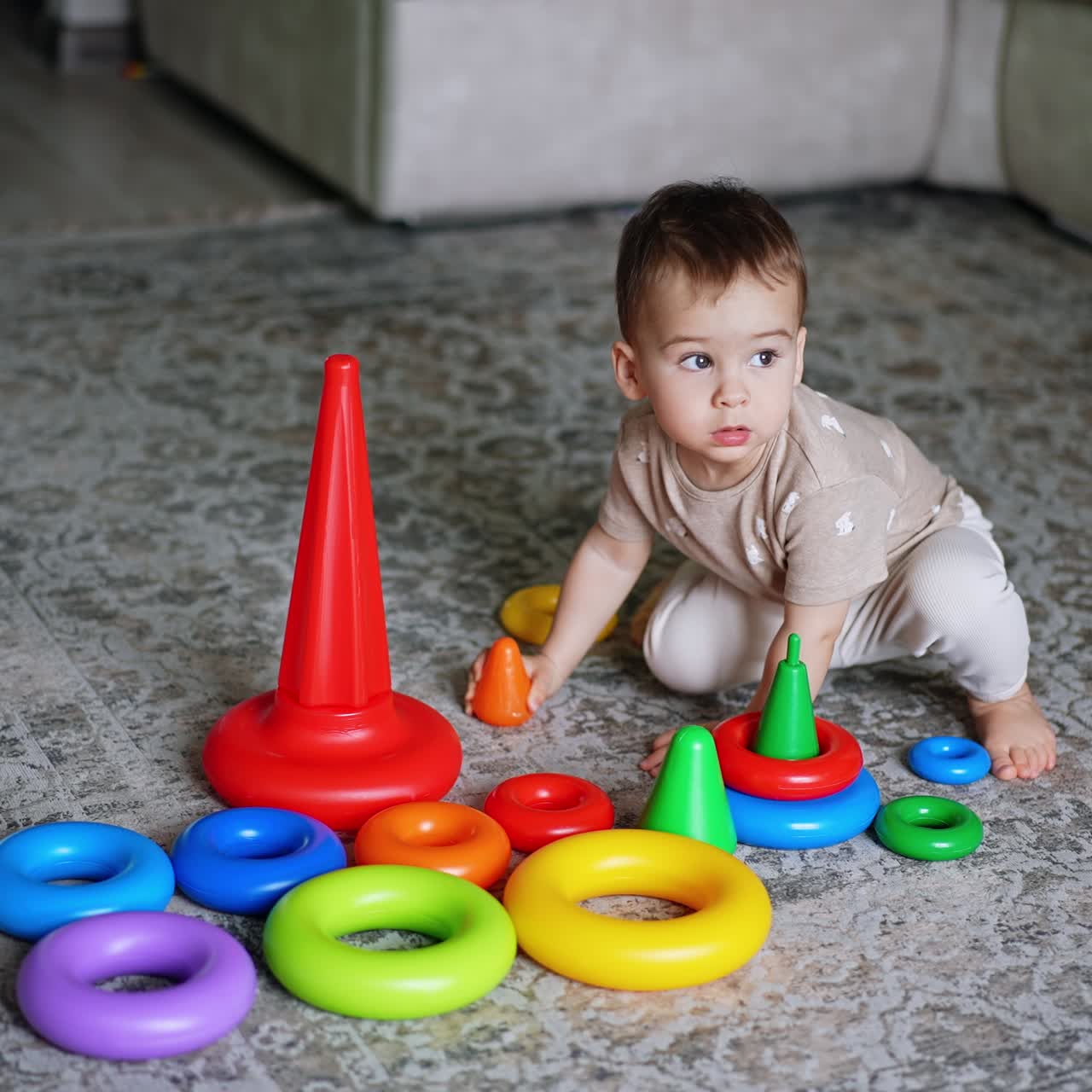 Cute toddler sitting on the floor holding a piece from toy pyramid. Baby boy notices something, stands up to go there
