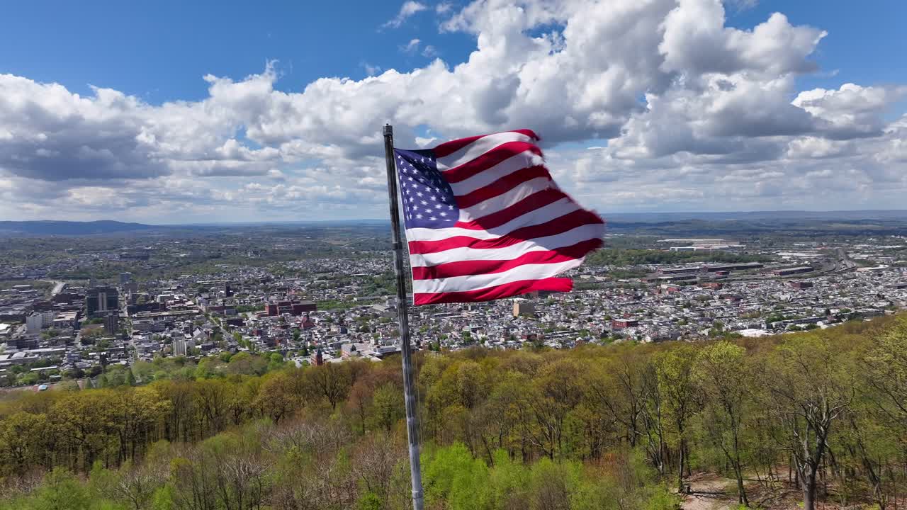 imágenes aéreas de una bandera estadounidense ondeando en la cima de una colina con vistas a un extenso paisaje urbano y bosques verdes exuberantes bajo un cielo azul brillante con nubes dispersas