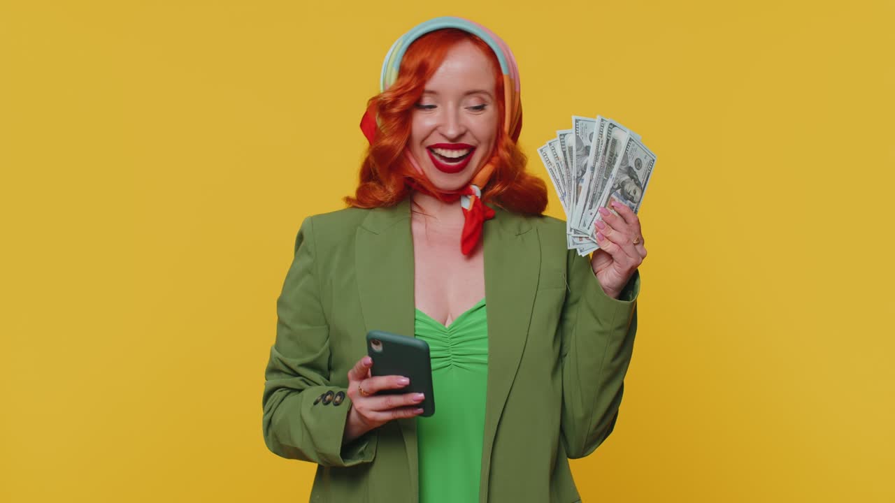 Donna dai capelli rossi felice che guarda lo schermo dello smartphone rallegrandosi sinceramente per una vittoria, ricevendo denaro contante in dollari.
