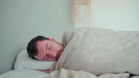 hombre dormido, blanco caucásico con barba, se despierta por la mañana, se estira cansado, abre los ojos, se levanta relajado en el interior del dormitorio