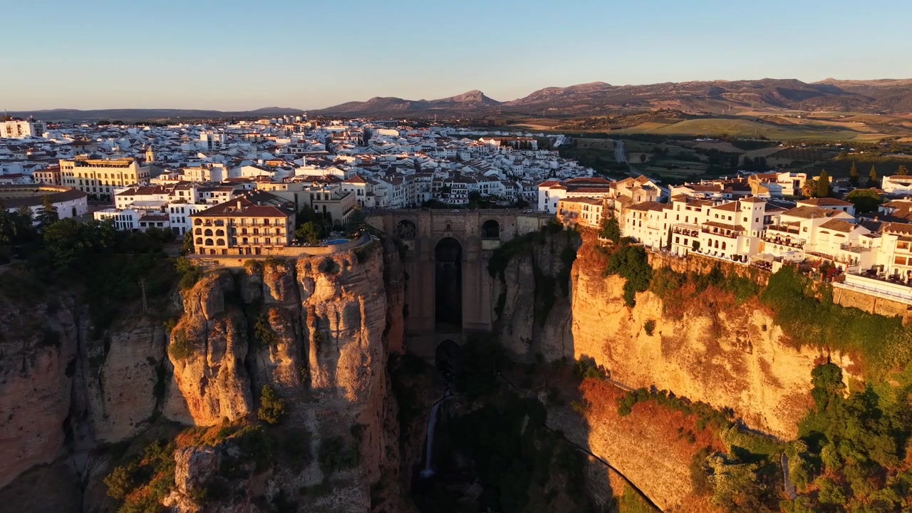 Wide drone aerial view of Puente Nuevo Bridge and the historic whitewashed town of Ronda, Spain during golden hour with dramatic cliffs and surrounding countryside