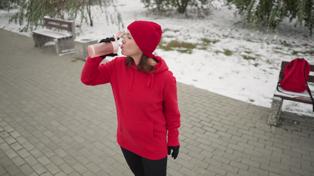 dama con capucha roja y gorra bebiendo de una botella de agua rosa mientras está de pie al aire libre en un parque cubierto de nieve, rodeada de árboles, una atmósfera de invierno serena y una bolsa roja colocada en un banco cercano