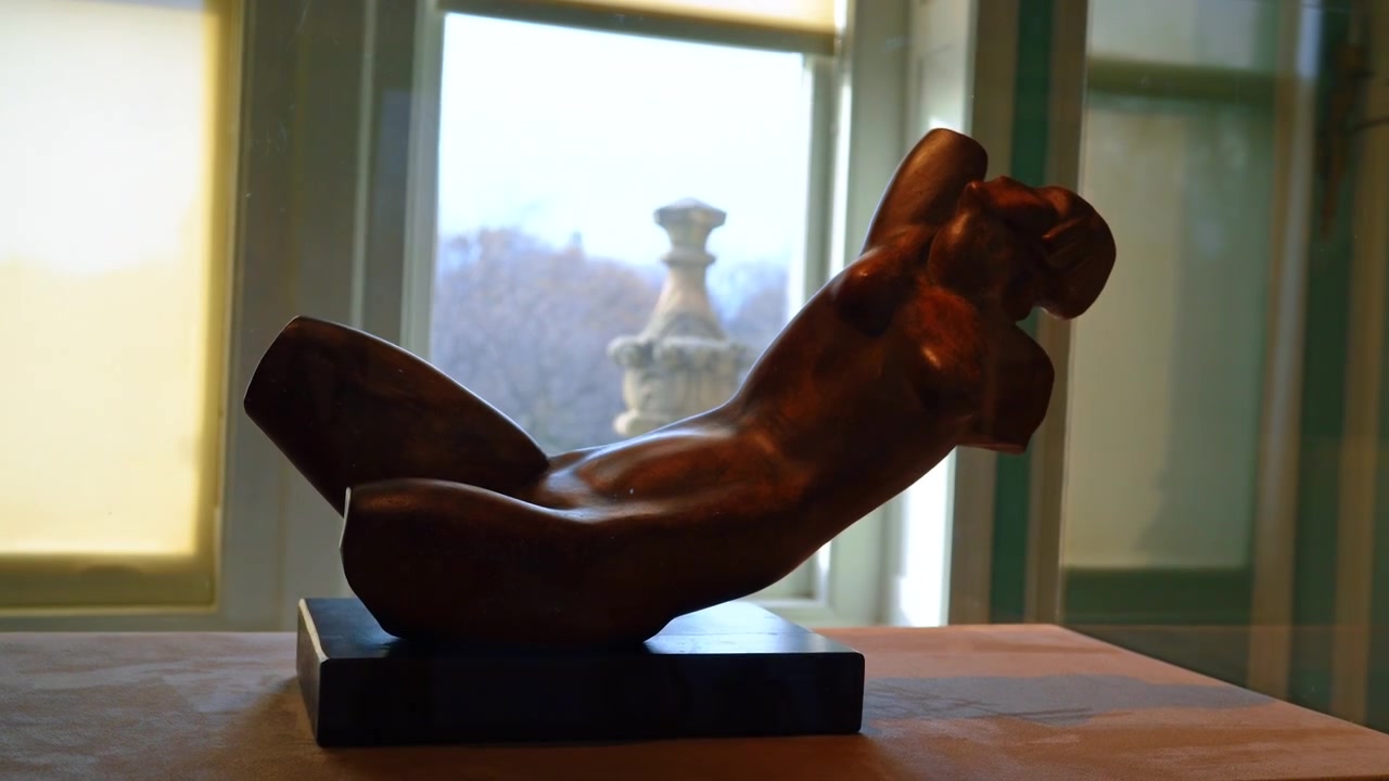 Scultura astratta di torso in bronzo su piedistallo vicino a una finestra, New York. Vista ravvicinata di una figura reclinata, base scura, luce laterale calda, dettaglio architettonico sfocato all'esterno.