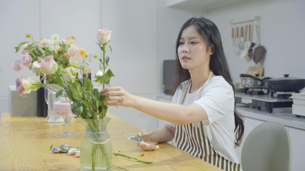 Konsep Florist: Wanita Muda Tersenyum Merangkai Buket Bunga Segar Berwarna-warni di Vas Kaca Dapur Modern, Menciptakan Komposisi Bunga Cerah untuk Ruang Keluarga