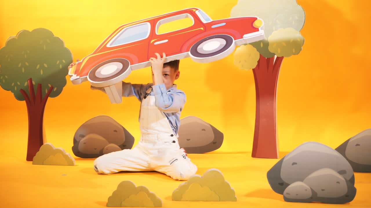 Cheerful little boy plays in the studio with toyful scenery on the yellow background. Happy kid with toyful carton car in his hands.