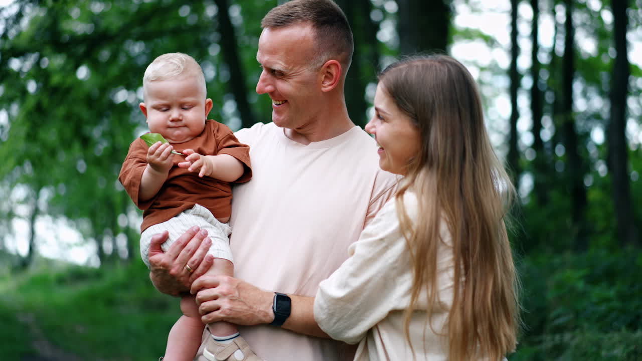 Happy Caucasian family with a baby of one year old stand in the nature. Lovely child holds a green leaf and gives it to his mom to smell.