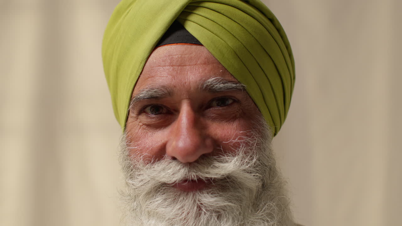 retrato de estudio de cerca de un hombre sikh mayor sonriente con barba y turbante contra un fondo plano
