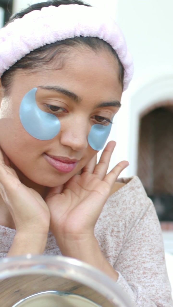 Vertical video: Glancing in mirror, woman checking blue eye patches at home with pink headband