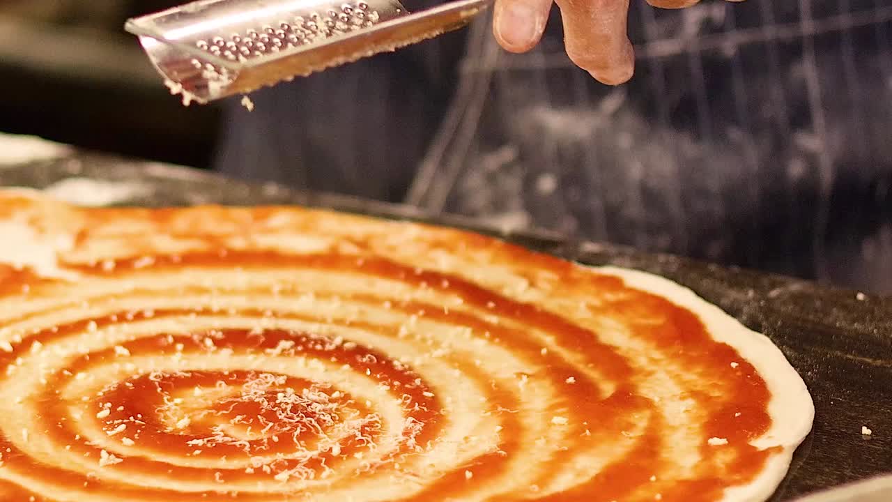 Close-up of cheese being grated over a tomato sauce spiral on pizza dough.