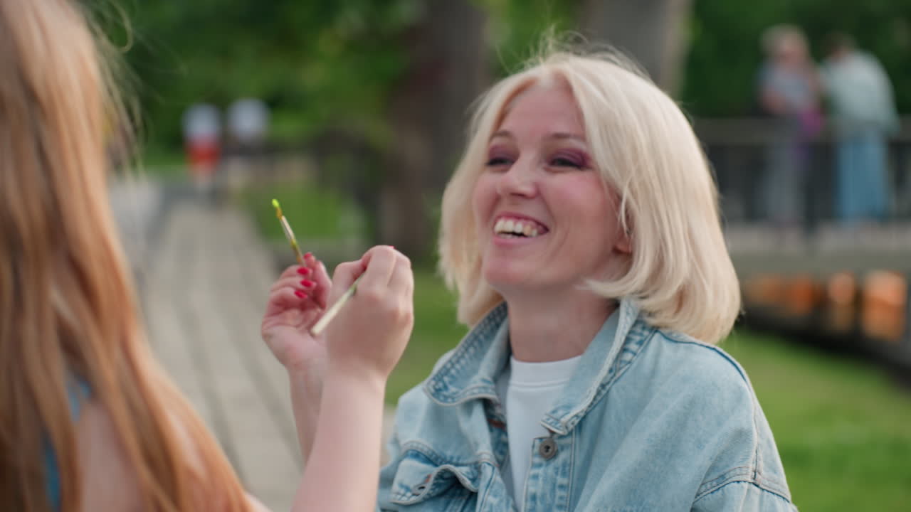 close up elegant mother and daughter face to face playfully touching cheeks with paint brush while seated outdoors by water against blurred green foliage in warm evening light with smiles