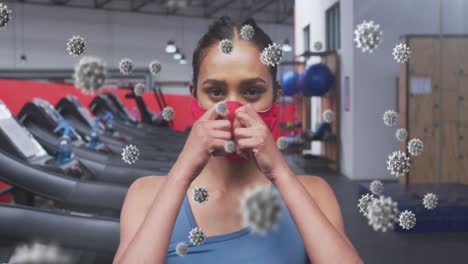 multiple covid-19 cells floating against portrait of woman wearing face mask at the gym