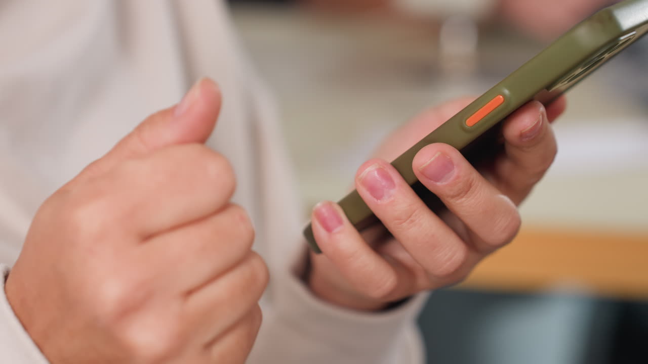 Close up of customer hands holding green smartphone with orange button carefully browsing screen with index finger during casual interaction in modern workspace wearing light colored outfit indoors