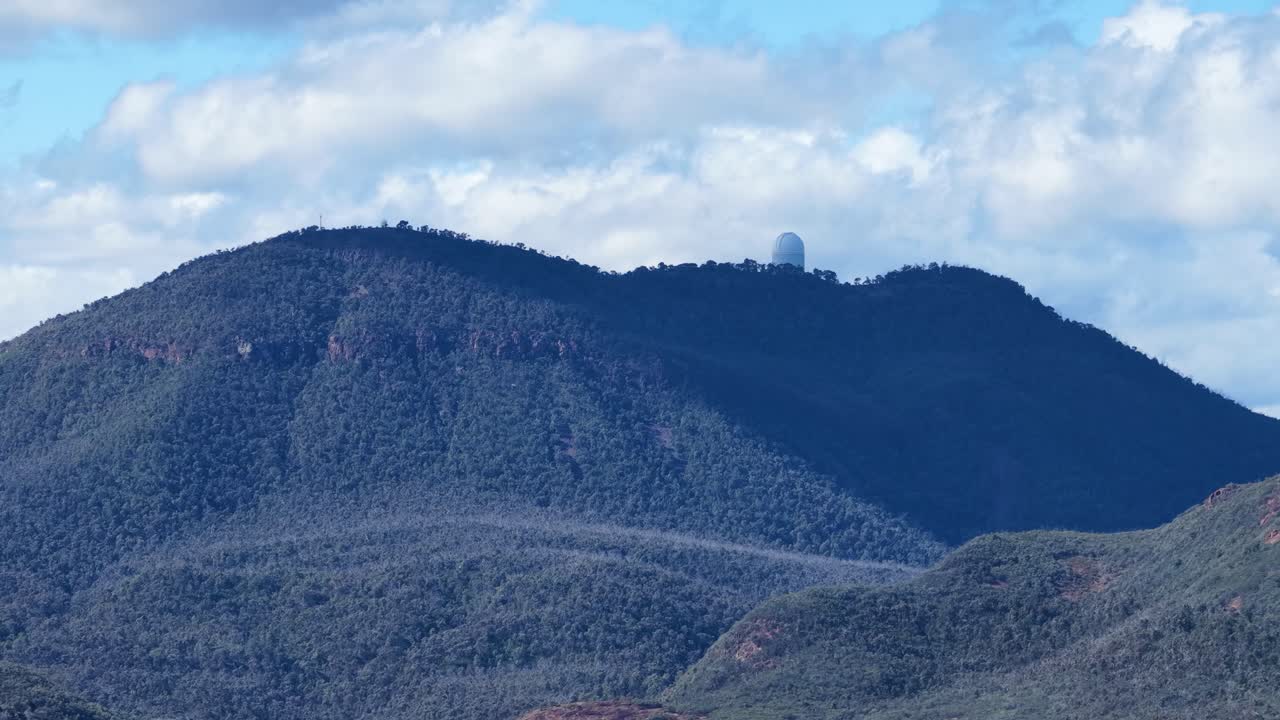 A large observatory dome slowly rotates atop a forested mountain ridge under bright daylight, with scattered clouds and clear atmospheric conditions