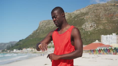 Hombre-Afroamericano-Revisando-Su-Reloj-Inteligente,-Tomando-Un-Descanso-En-El-Ejercicio-Al-Aire-Libre-Junto-Al-Mar