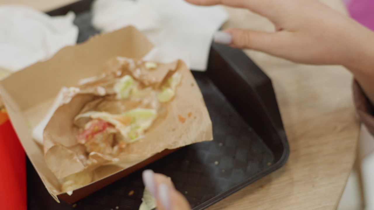 Persona che si pulisce le dita dai condimenti, pasto al chiuso con lattuga e salsa sparse, scena informale al chiuso con tovagliolo per pulire le macchie di hamburger dalle dita e avanzi sparsi