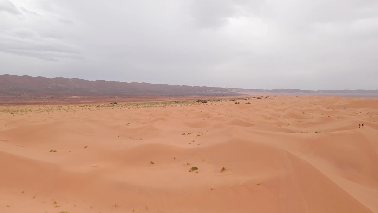 Drone shot over small sandy dunes with big mountains behind in morocco desert during cloudy day