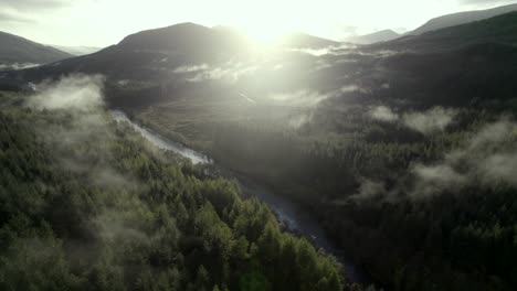 Drohnenaufnahmen-Aus-Der-Luft-Erheben-Sich-Langsam-über-Einem-Fluss-Und-Einem-Wald-Aus-Nadelbäumen,-Während-Lichtstrahlen-Tief-Hängende-Wolken-In-Den-Baumwipfeln-Beleuchten-Und-Die-Sonne-Hinter-Bergen-Untergeht