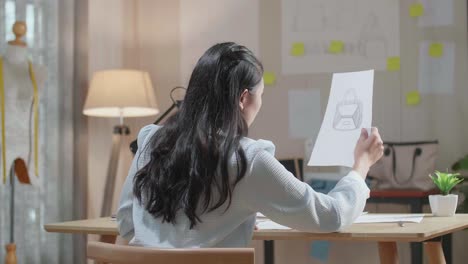 아시아 여성 디자이너의 뒷면은 손 가방을 보면서 머리를 긍정적으로 흔들고 손으로 레이아웃을 스케치합니다.