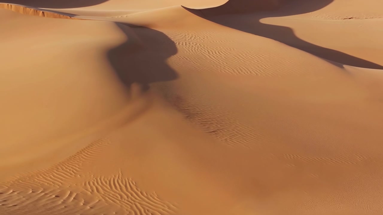 Vista dall'alto di dune di sabbia desertiche illuminate dal sole che riempiono l'inquadratura; una grande duna centrale con creste affilate e pendii lisci mostra creste scolpite dal vento, a forma di mezzaluna