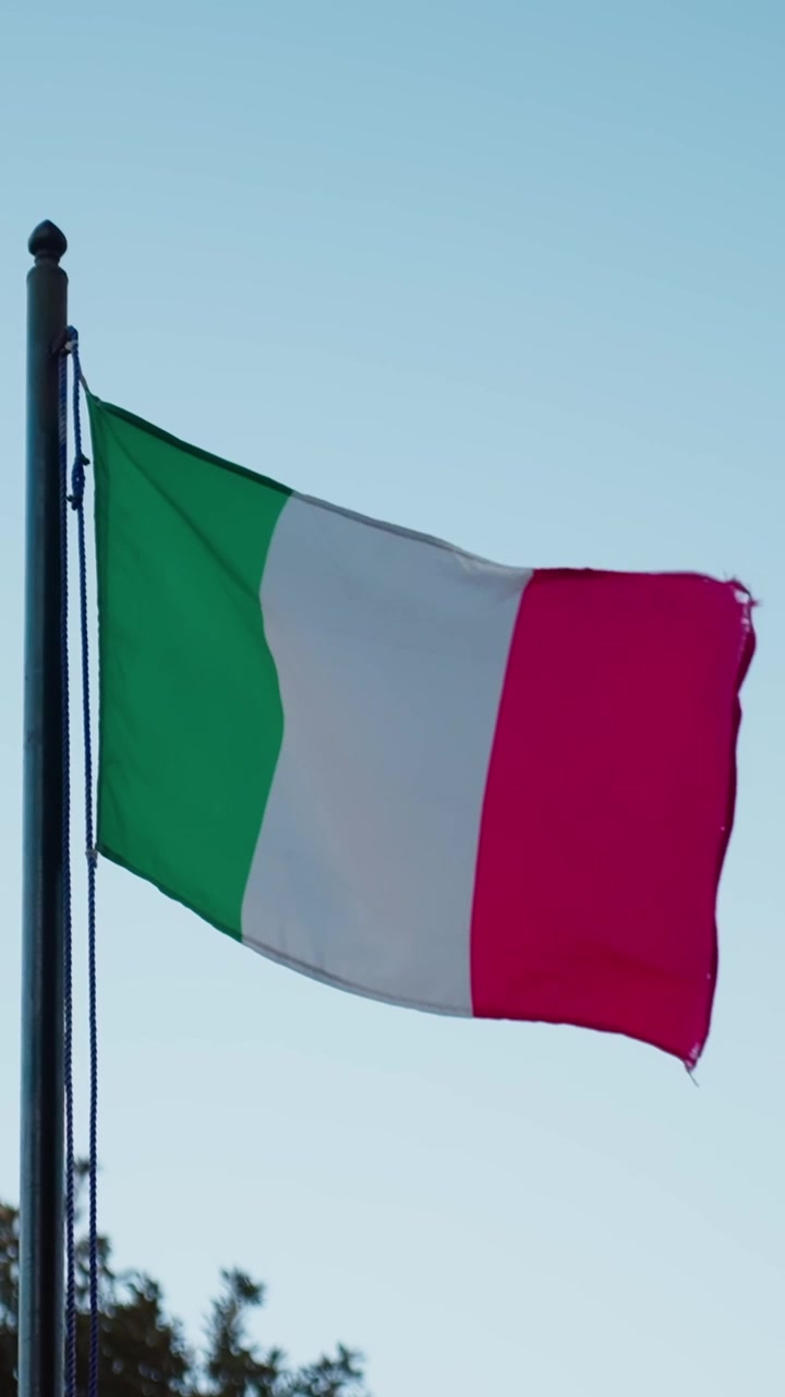 Bandiera italiana che sventola su un'asta contro un cielo azzurro limpido, con il tessuto verde, bianco e rosso che si muove nella brezza in una semplice inquadratura verticale patriottica