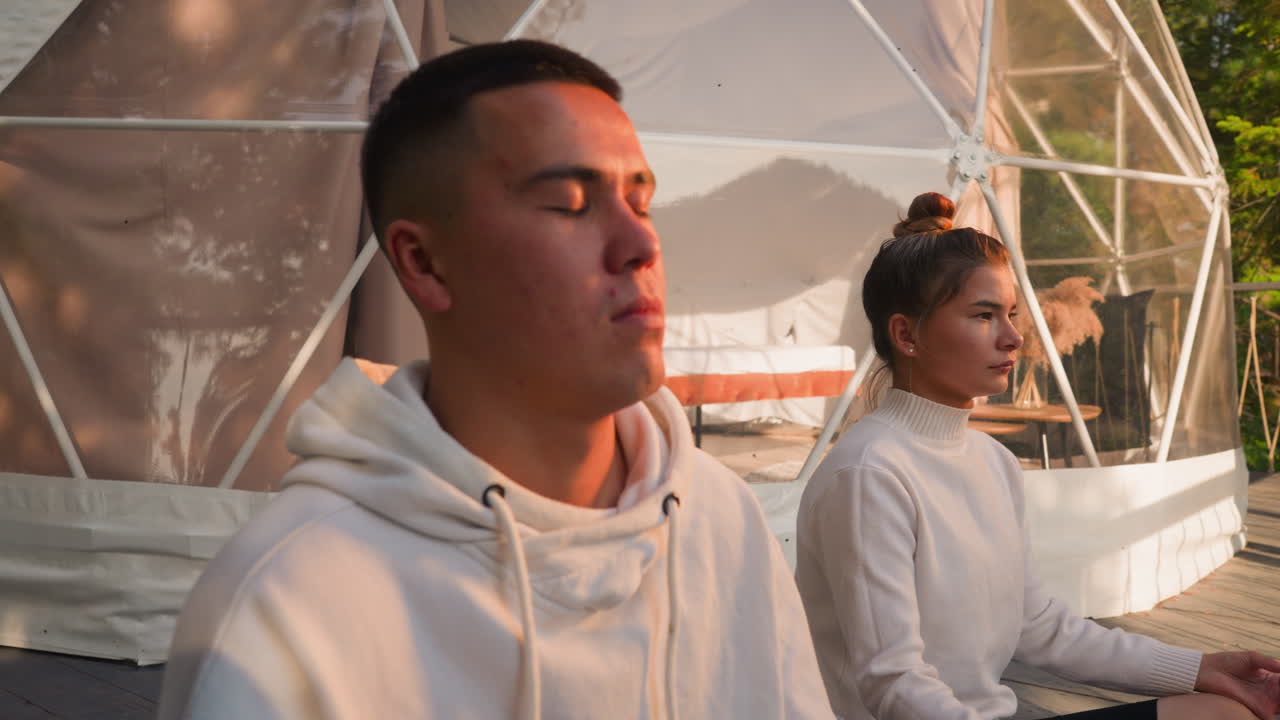 una pareja joven medita descansando en glamping de cerca. un hombre y una mujer atentos hacen prácticas relajantes en un eco-resort al atardecer. vacaciones en la naturaleza salvaje con comodidad