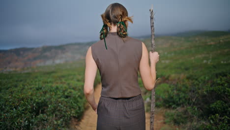 woman journeying green hill with stick alone. rear view serene girl walking path