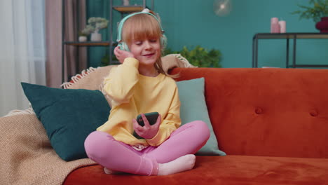 child girl kid with smartphone in headphones dancing while listening music at home alone on sofa