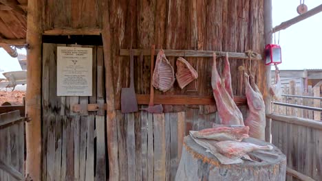una carnicería rústica con carnes y herramientas colgantes, ubicada en un entorno histórico al aire libre con estructuras de madera