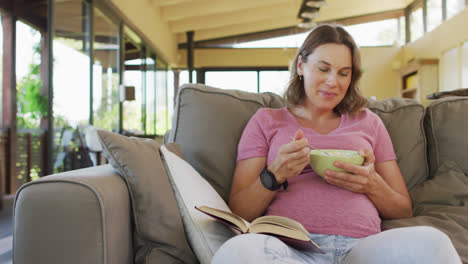 Mujer-Embarazada-Caucásica-Relajada-Tumbada-En-El-Sofá,-Comiendo-Y-Leyendo-Un-Libro