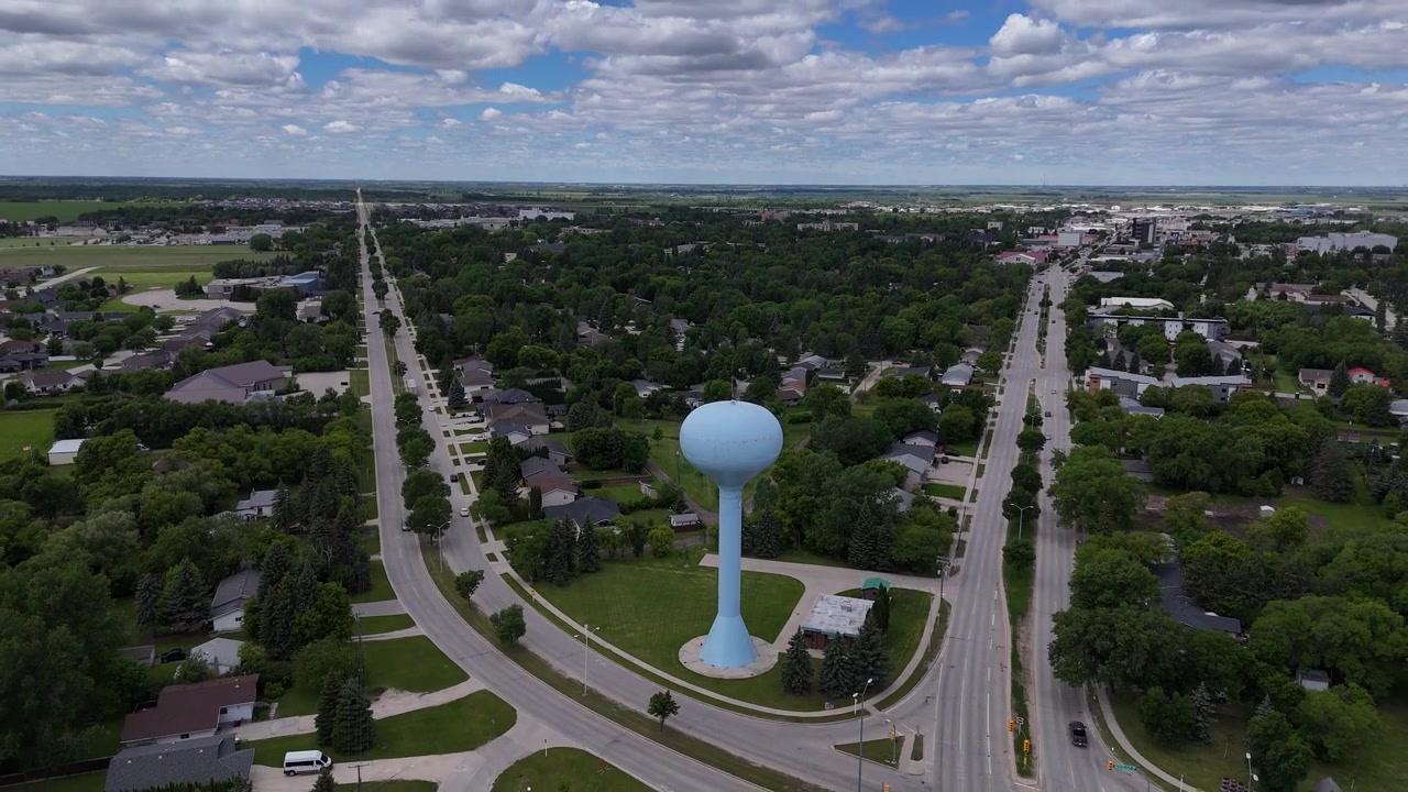 Drone bevæger sig op og væk fra det lyseblå vandtårn i Steinbach, Manitoba, og afslører den hævede tank, veje, biler, huse, træer, forstæder og grønt græs om sommeren