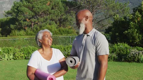 Portrait-of-african-american-senior-couple-holding-yoga-mats-smiling-while-standing-in-the-garden