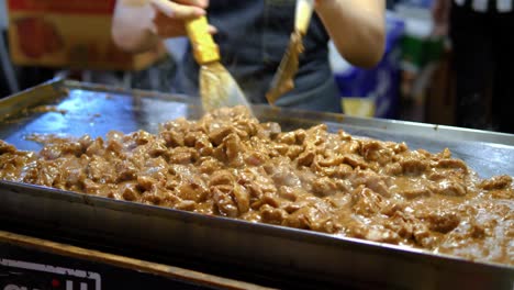 Chef-Revolviendo-Estofado-De-Carne-En-Una-Parrilla-Plana-Abierta,-Mercado-Nocturno-De-Tren-Bangkok