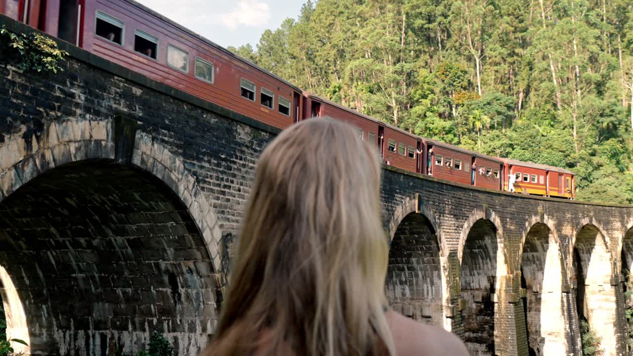 A young woman stands at a viewpoint near the Nine Arch Bridge in Ella, Sri Lanka, gazing at a colorful train as it moves across the historic stone arches.