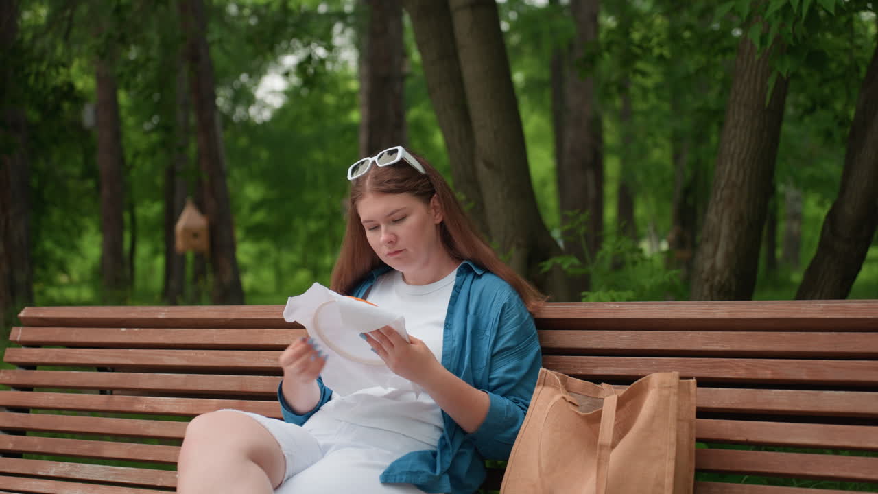 Young student sitting on wooden park bench embroidering fabric with needle under soft summer light, wearing blue shirt and white outfit, concentrating calmly on creative needlework