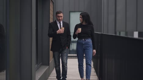 portrait of confident male and female coworkers walking in slow motion talking. handsome caucasian man and beautiful woman discussing startup strategy idea on break strolling with coffee cups.
