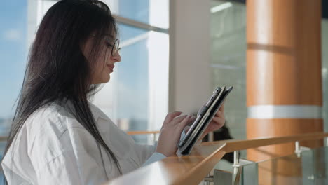 side view of woman in white shirt using tablet with joyful expression in bright indoor space featuring wooden columns, glass railings, and soft daylight through large windows