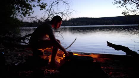 joven cocinando su cena en el fuego del campamento junto al lago al anochecer