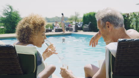 retired senior couple on loungers relaxing by swimming pool on summer vacation drinking champagne