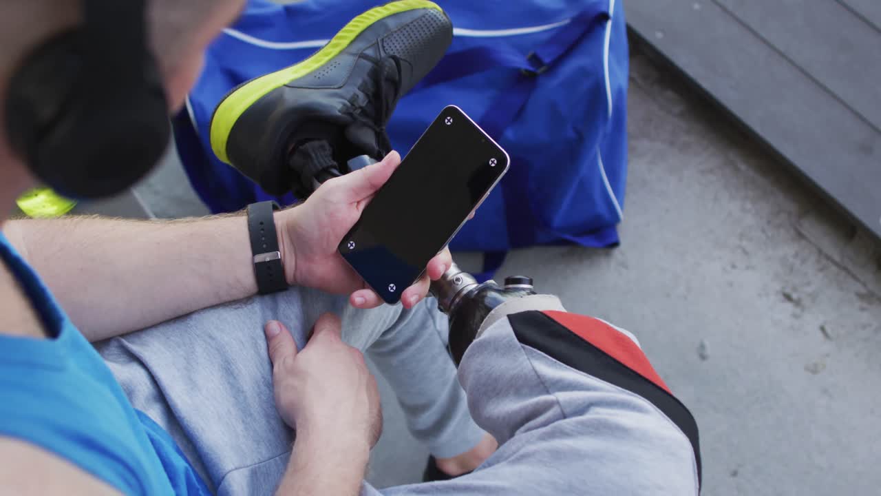 atleta masculino caucásico discapacitado con una pierna protésica sentado, con auriculares, usando un teléfono inteligente