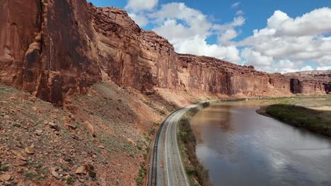 drone flying over curved highway in the desert, in between tall red cliffs, railroad tracks and river, blue sky with clouds early spring, potash road in moab utah