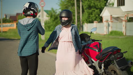 two friends stand near a parked power bike, the woman in pink playfully moves her gown while the other touches her chin in playfully,in a peaceful residential area