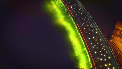 Aerial-top-down-view-flying-forward-over-Starlight-Bridge-or-Anh-Sao-Bridge-at-night,-a-pedestrian-bridge-with-strong-colored-lights-and-waterfall-in-District-7-of-Ho-Chi-Minh-City-or-Saigon,-Vietnam