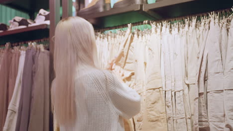 back view of shopper browsing rack of denim jeans in clothing store, pulling pair from hanger clips while boots displayed on top shelf under warm boutique lights with blurred racks behind