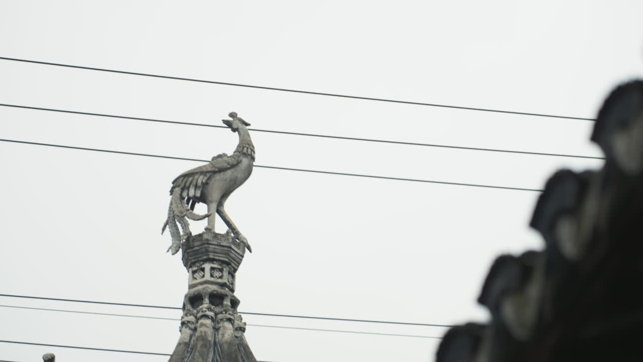 Udsmykket føniksstatue på tagryggen mod himlen, vejrbidt metalkam placeret på tempelspir, diagonal tag-silhuet indrammer højre side, lagdelte elledninger skærer igennem scenen, overskyet stemning med dæmpede farver