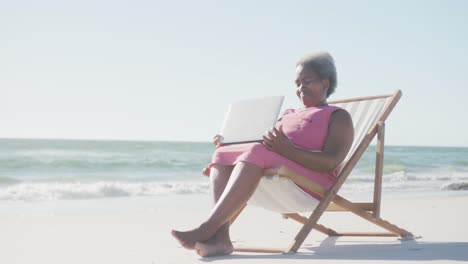 Glückliche-ältere-Afroamerikanische-Frau,-Die-Auf-Einem-Liegestuhl-Sitzt-Und-Am-Strand-Einen-Laptop-Benutzt,-In-Zeitlupe